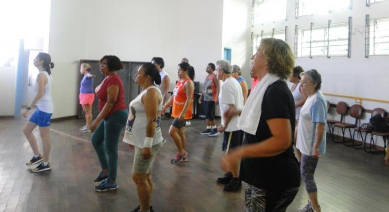 Dez cidadãos na Academia da Cidade, fazendo ginástica, em uma sala, durante o dia. 