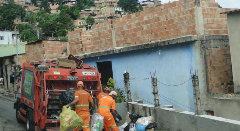  Caminhão que recolhe lixo ao lado de casa, neste local, três garis recolhem o lixo.