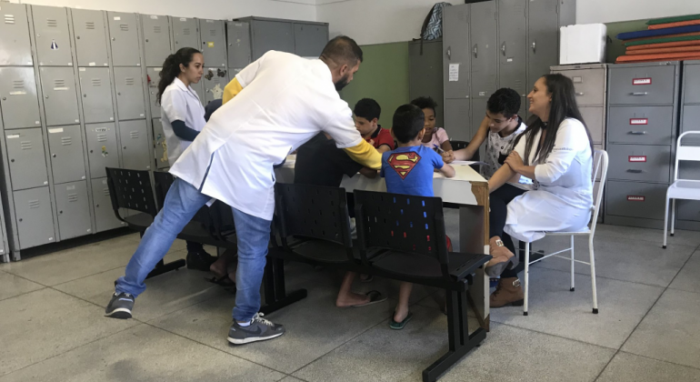 Grupo Aprender promove a potencialização de práticas que contribuam para melhorias no processo de aprendizagem de crianças e adolescentes. TRês crianças realizam atividades em mesa, acompanhadas por orientadores.
