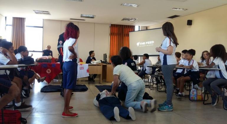 Estudante simula exercício de massagem cardíaca, acompanhada por duas estudantes de pé e mais de quinze sentadas, em uma sala. 