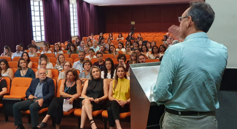 Projeto Bem-Vindo, implantado nas unidades de saúde da capital para melhorar o atendimento, apresenta resultados positivos Homem, no palco, fala para pessoas sentadas em um auditório