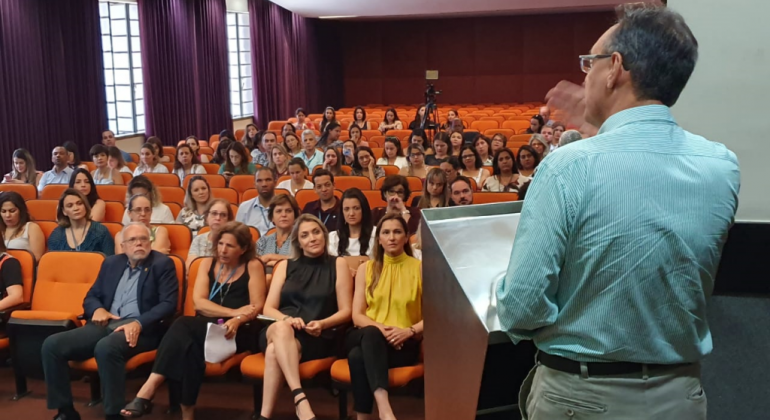 Homem, no palco, fala para pessoas sentadas em um auditório