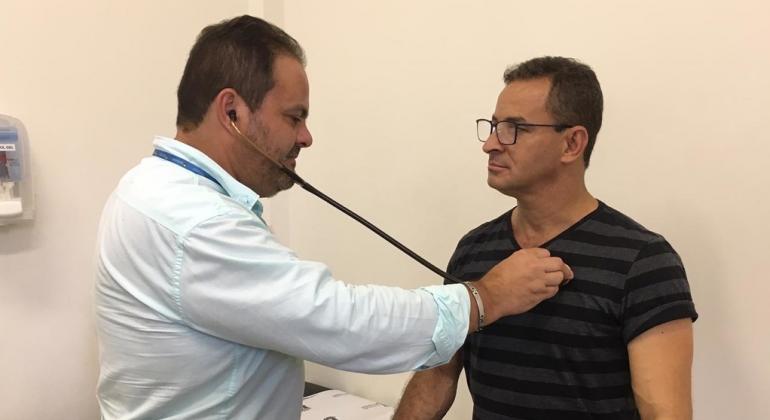 Homem confere os batimentos cardíacos de paciente com estetoscópio, em sala de centro de saúde. 