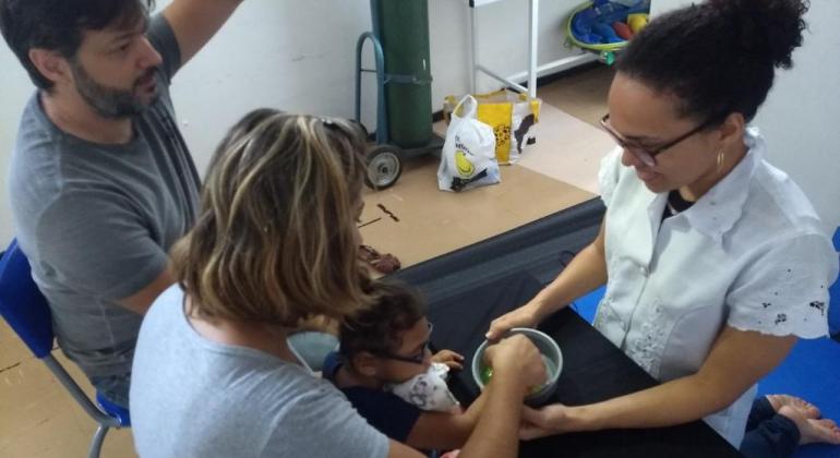 Pais acompanham filha com óculos, sentada no colo da mãe, que aprende técnica de habilitação visual. 