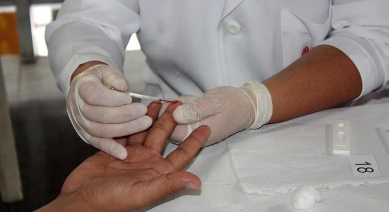 Mãos enluvadas e de jaleco branco encostam fita de testagem em um dedo da mão do cidadão. 