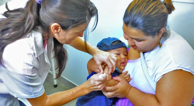 Os Centros de Saúde de BH estarão abertos das 8h às 17h no dia 19/10 para vacinar crianças de seis meses a menores de cinco anos contra o sarampo. Foto: Divulgação PBH Criança, no colo da mãe, recebe vacina por uma enfermeira