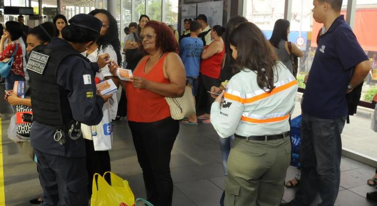 Guarda Municipal e agente da BHTrans distribuem folhetos educativos e apito sobre prevenção a assédio sexual para duas mulheres. 