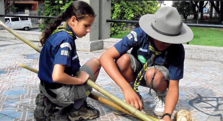 Escotismo da Guarda Municipal Duas crianças, uma menina e um menino, manuseiam bambus