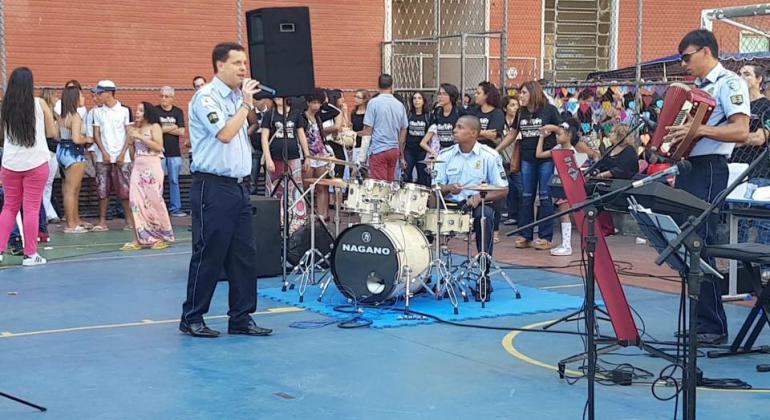 Cantor e baterista da Banda da Guarda Municipal se apresenta em quadra esportiva; ao fundo, plateia com mais de 60 pessoas. 