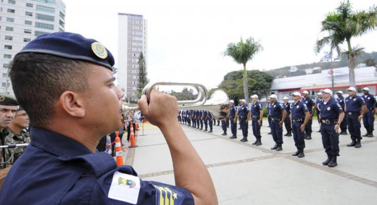 Guarda Municipal toca instrumento de sopro enquanto cerca de vinte guardas desfilam, durante o dia. 