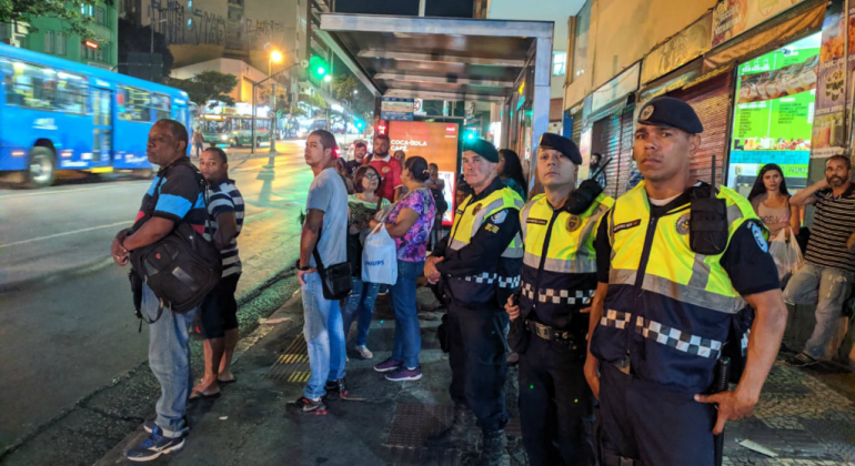 A Guarda Civil Municipal selecionou dez pontos de ônibus para implantar a novidade, que visa potencializar a Operação Viagem Segura. Foto: Divulgação PBH Três guardas municipais ao lado de um ponto de ônibus com mais de seis pessoas, à noite.