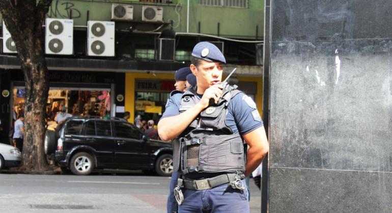 Guarda municipal, em esquina de uma rua, fala com rádio preso ao ao ombro, durante o dia.