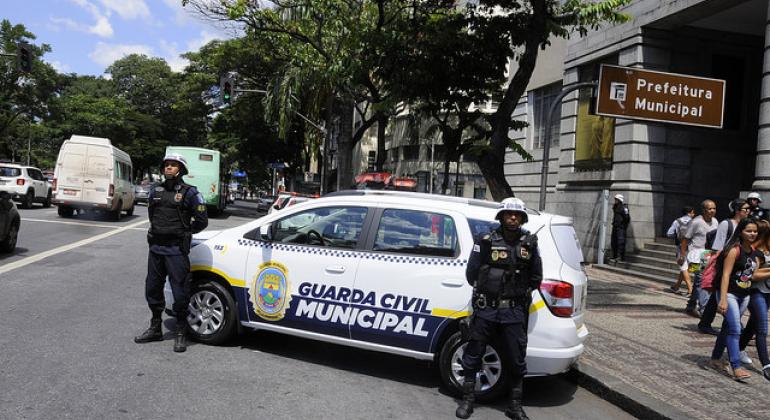 Dois Guardas civis municipais, com capacetes, à frente de um carro da Guarda, na frente da Prefeitura de Belo Horizonte, durante o dia. 