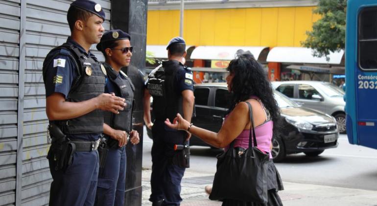 Guarda Municipal destaca a importância de que cada cidadão contribua para garantir uma folia com segurança. Foto: Divulgação PBH Foliã de Carnaval conversa com guarda municipal feninina, ao lado de guarda masculino, durante o dia. Outro guarda está ao fundo.
