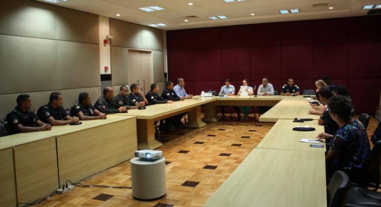 Dezessete pessoas em reunião em torno de uma mesa em forma de "u".