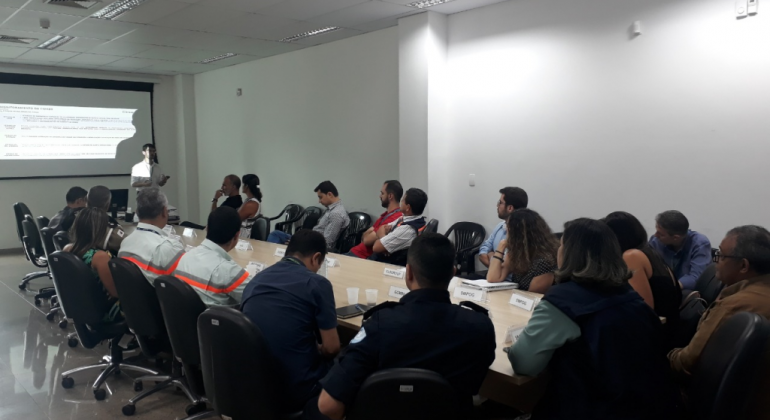Representantes do COP-BH e da empresa Elogroup reuniram-se, no dia 6/11, para discutir o fim da consultoria prestada à Prefeitura de BH. Foto: Divulgação PBH Cerca de dezessete pessoas, sentadas, reunidas em torno de uma mesa, e um homem em pé que palestraao lado de uma projeção na parede.
