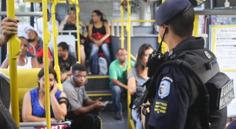 Guarda municipal de costas, dentro de ônibus com passageiros.