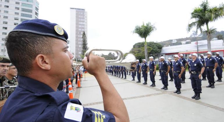 Guarda civil municipal toca instrumento de sopro; à frente, mais de trinta guardas enfileirados. 