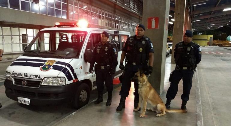 Três guardas municipais e um cão ao lado de uma van da Guarda Municipal, à noite. 