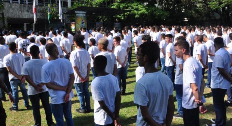 Mais de duzentos jovens, todos de camiseta branca e mãos para trás, observam a bandeira, durante o dia, a céu aberto. 