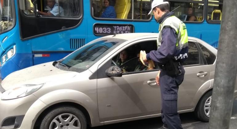 Guarda municipal entrega folder educativo a motorista de carro particular. Um ônibus aparece ao fundo da imagem