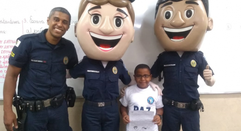 Palestras sobre respeito e sensibilidade promovidas pela Patrulha Escolar mudam rotina de alunos em escola do Barreiro. Foto: Divulgação PBH Guarda Municipal, estudante e dois guardas com máscaras humanas de guarda municipal similares a desenho animado, posam para foto.