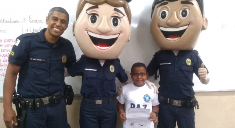 Guarda Municipal, estudante e dois guardas com máscaras humanas de guarda municipal similares a desenho animado, posam para foto. 