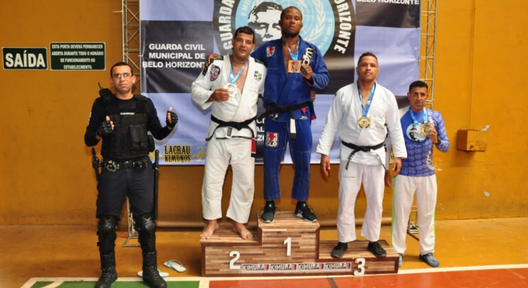 2º Campeonato de Jiu-jitsu, realizado no dia 3/11, na quadra da E. M. Marconi, no Gutierrez, superou a marca de 100 atletas inscritos. Foto: Divulgação PBH Pódio com três campeões do campeonado de Jiu-jitsu, com dois homens ao lado.