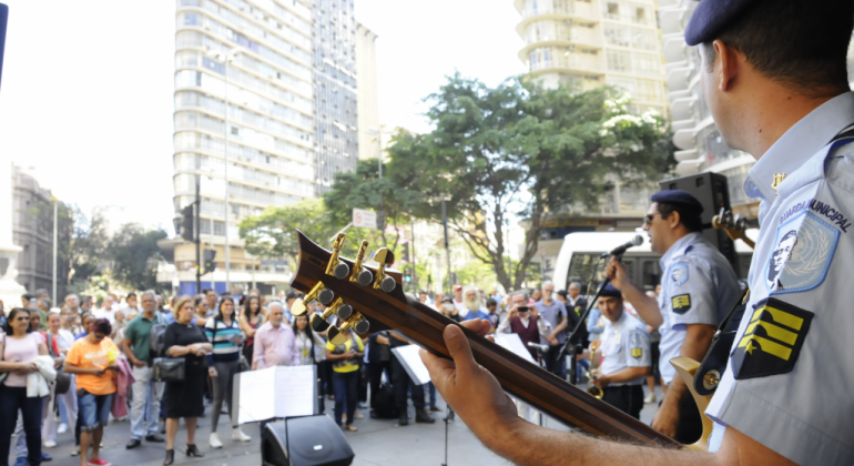 A proposta do grupamento é levar a música a todas as regionais de Belo Horizonte ao longo do ano. Foto: Divulgação PBH Quatro integrantes da banda da Guarda Municipal se apresentam à direita, imagem parcial da banda dá espaço ao grande público, de pé, na Praça Sete, ao fundo, à esquerda.