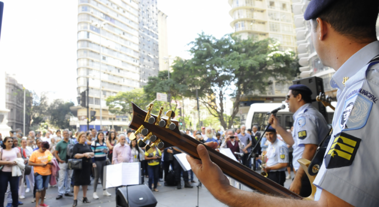 Quatro integrantes da banda da Guarda Municipal se apresentam à direita, imagem parcial da banda dá espaço ao grande público, de pé, na Praça Sete, ao fundo, à esquerda.