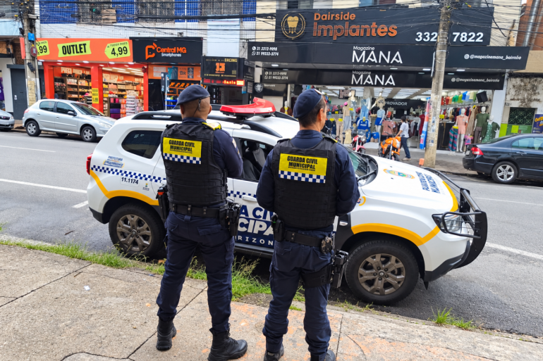 Barreiro + Seguro Dois agentes da Guarda Civil Municipal de Belo Horizonte, vistos de costas, estão em uma calçada ao lado de uma viatura da corporação estacionada em uma via. Ao fundo, há estabelecimentos comerciais, veículos estacionados e pedestres.