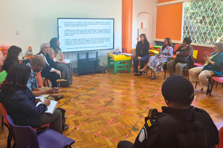 Grupo de Trabalho Segurança nas Escolas Pessoas sentadas em círculo numa sala de reuniões assistem a uma apresentação em tela sobre prevenção da violência contra crianças.