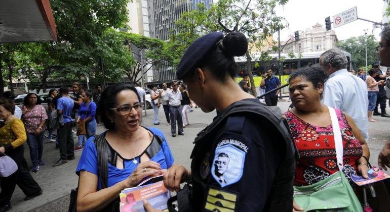 Grupo contra o Assédio Sexual da Prefeitura estará na Praça da Liberdade com apitos e folhetos educativos na quinta-feira, dia 20/12. Foto: Rodrigo Clemente/PBH Guarda municipal feminina aborda cidadã na Praça Sete com material informativo. Foto ilustrativa.