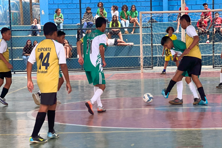 Copinha 2025 Jovens jogam futebol em quadra coberta enquanto público assiste na arquibancada ao fundo. Um garoto de verde disputa a bola perto do gol.