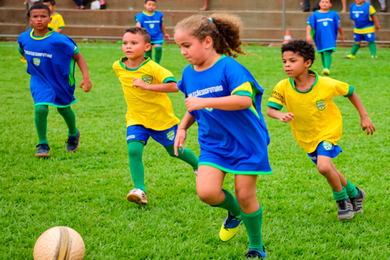  Foto mostra crianças jogando futebol em um campo de grama sintética, usando uniformes azuis e amarelos. Ao fundo, há outras crianças e arquibancadas, em um ambiente de esporte, diversão e convivência.