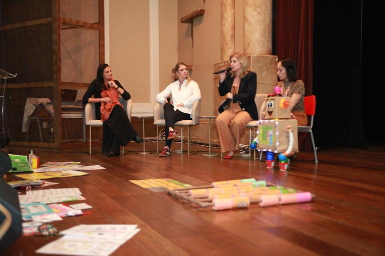 Quatro mulheres sentadas em cadeiras em uma sala de teatro com trabalhos infantis expostos no chão na parte da frente, uma das mulheres está falando segurando um microfone. 