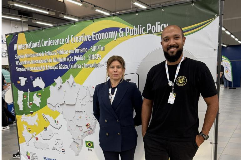 Débora Giordana, gerente de Promoção Turística da Belotur, e Márcio Ribeiro, gerente de Inovação Turística da Belotur, posam para foto durante o Seminário Internacional de Pesquisa em Políticas Públicas de Turismo, realizado em Viçosa. Ao fundo, banner apresenta a programação do evento.