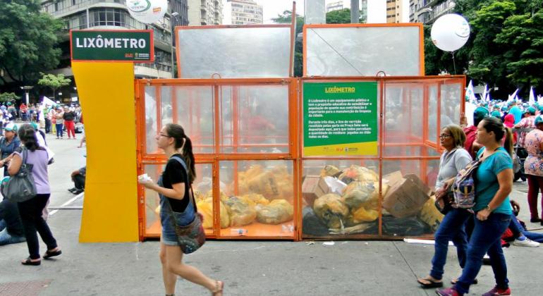 Quatro recipientes transparentes, cada um medindo 2m², estarão de volta ao centro de BH para armazenar os resíduos de varrição recolhidos. Foto: Jefferson Andrade Na Praça Sete de Setembro, um grande recipente laranja com grade escrito "Lixômetro" armazena sacos com o lixo recolhido na região. Pessoas transitam ao lado do lixômetro.