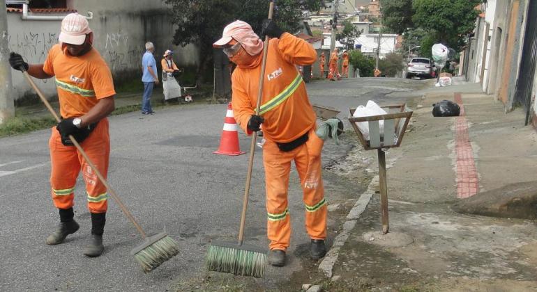 Dois garis da SLU varrem grama de capina realizada, em uma rua de bairro de Belo Horizonte, durante o dia. 