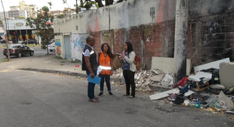 Três pessoas usando colete conversam ao lado de lixo irregular 