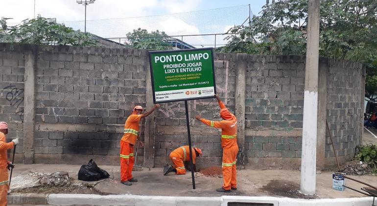 Enquanto um tecnico da SLU varre a rua, outros três implantam a placa de Ponto Limpo em uma calçada, durante o dia. 