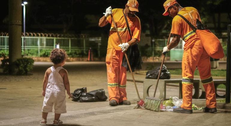 Criança observa dois garis varrerem o chão, em local próximo a banco de cimento. 