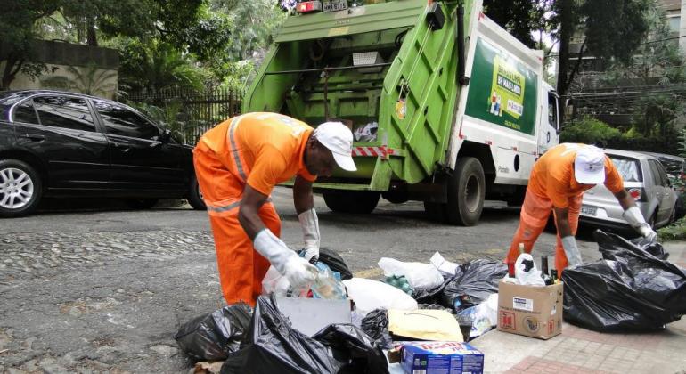 Dos garis da Prefeitura recolhem lixo reciclável na calçada, durante o dia. ao fundo, caminhão de lixo reciclável.
