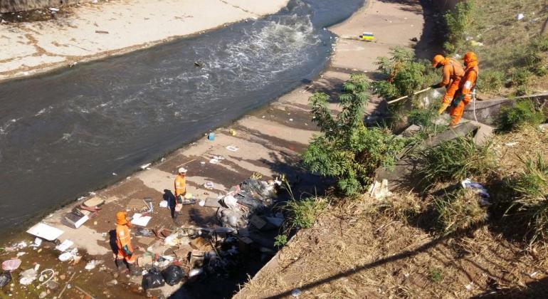 Dois técnicos da SLU realizam limpeza nas laterais do rio Arrudas, durante o dia. 
