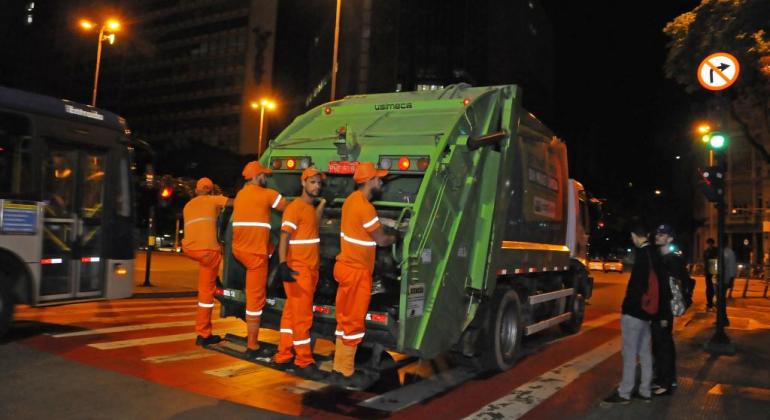 Toda a frota da SLU é rastreada por meio de GPS e, nos grandes corredores viários de BH, o serviço é executado seguindo cuidados especiais. Foto: Adão de Souza/PBH Quatro garis, de pé, da traseira de um caminhão de recolhimento de lixo da SLU, na praça Sete, à noite.