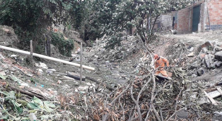 Funcionário da SLU, vestido de laranja, retira galhos de um córrego, durante o dia. 