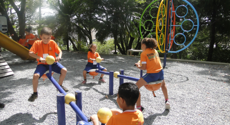 Quatro alunos da Rede Municipal de Educação, com camisetas laranja, brincam em brinquedos de uma praça, durante o dia. 