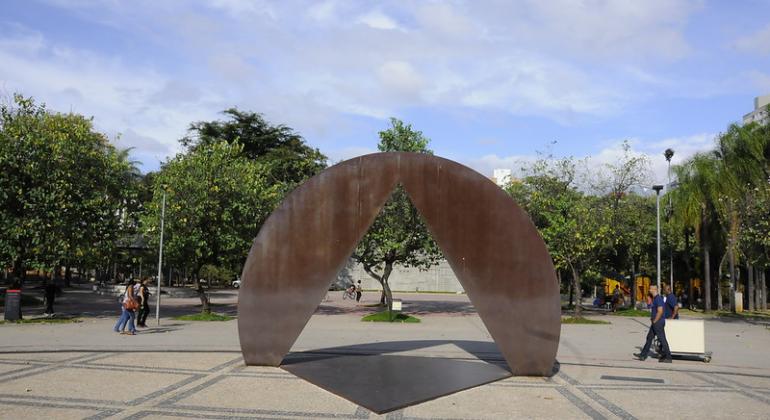 Praça da Assembleia, com escultura com o triângulo símbolo de Minas Gerais ao centro e pessoas passeando dos dois lados e muitas árvores ao fundo, durante o dia. 