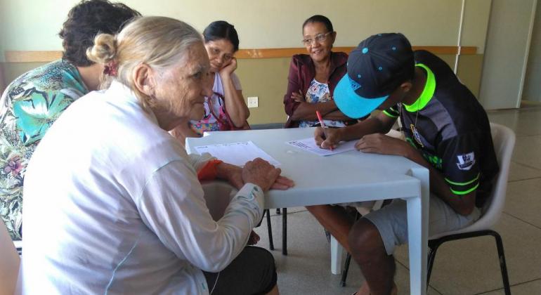 Vínculo intrafamiliar e comunitário é foco do trabalho de assistência social da Prefeitura de Belo Horizonte. Foto: Sandra Barros Idosa e mulher, sentadas à mesa, acompanham menor, também sentado, assinando um papel.