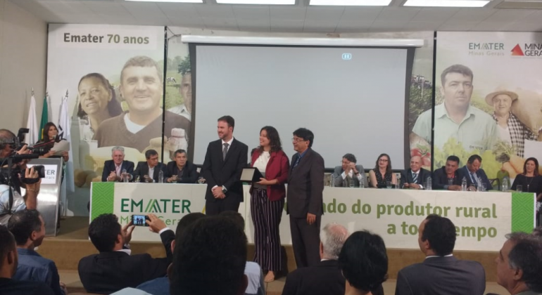 Emater homenageia política de segurança alimentar da Prefeitura Três pessoas de pé, em um palco. Uma delas, ao centro, é a Secretária Municipal de Assistência Social, Segurança Alimentar e Cidadania, segura uma placa de premiação